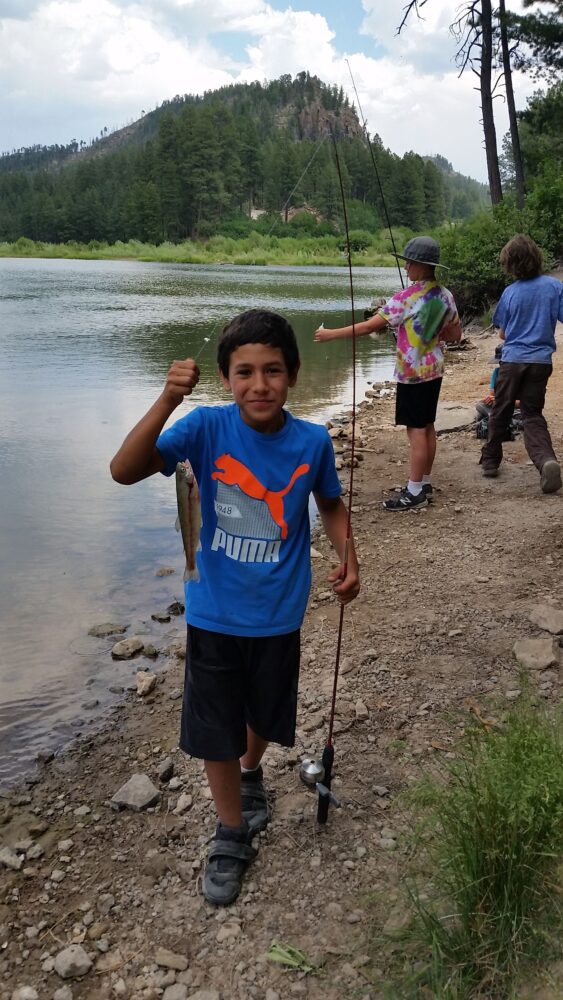 A boy in a blue shirt holds up a fish and fishing rod next to a body of water.